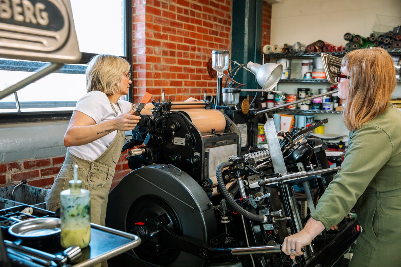 Two women working on a large printing press in a workshop.
Photo by Alyssa Broadus