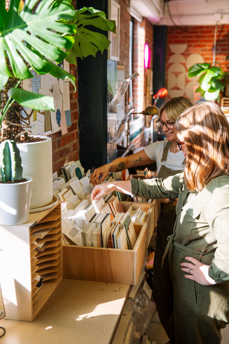 Two women in a casual setting, reaching into a box of files, with plants and office decor in the background.
photo by Alyssa Broadus