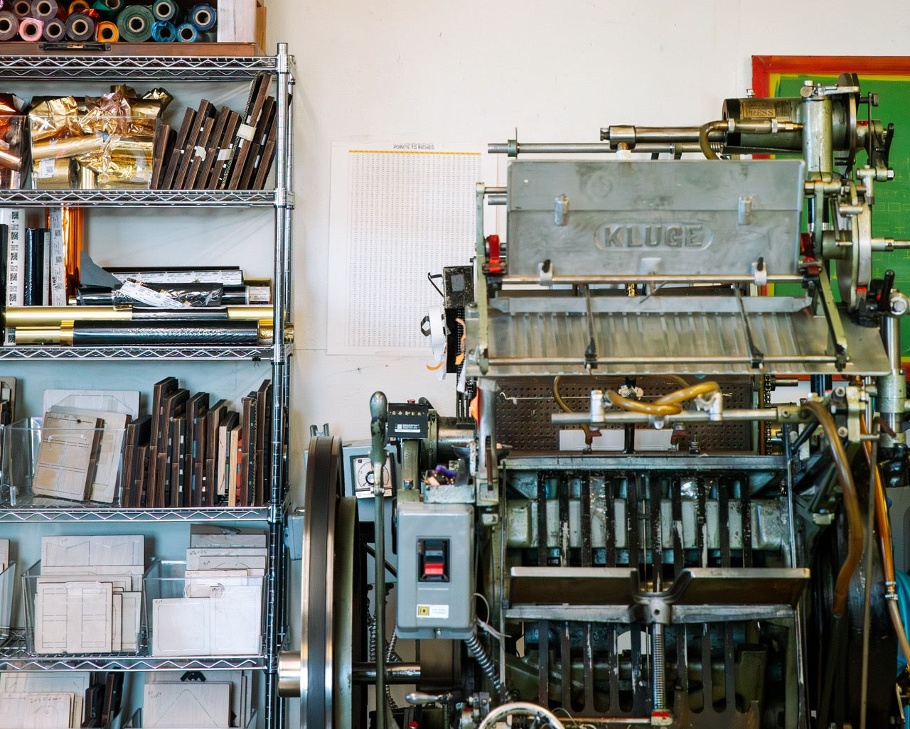 Printing press machine in a workshop with shelves of supplies
Photo by Alyssa Broadus